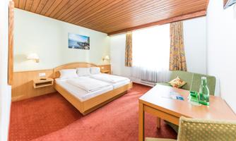 A cozy hotel room with a double bed, a table, and a chair. There is a large window with curtains and a wooden ceiling.