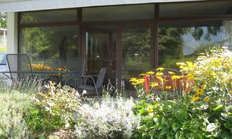 A cozy outdoor area with a table and chairs in front of a modern building. Colorful flowers and green greenery frame the porch.