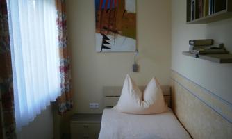 A simple bedroom with a bed, two pillows, and a nightstand. A modern artwork hangs on the wall, and a window with curtains lets in a lot of light.