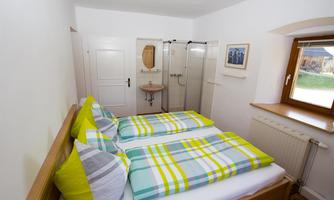 A cozy bedroom with two single beds and colorful bedding. The bathroom is separated by a glass door and there is a window with a view outside.
