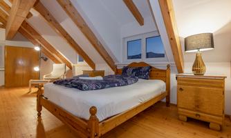 A cozy bedroom with a wooden ceiling and a large bed. Next to the bed is a bedside table with a lamp and a chair in the corner.