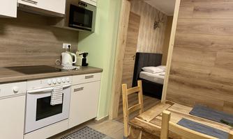 A modern kitchen with wooden accents and a dining table. In the background, a bedroom is visible.