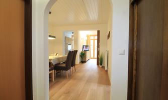 A bright and modern room with wooden floors. In the background, you can see a dining table with chairs and an open kitchen.