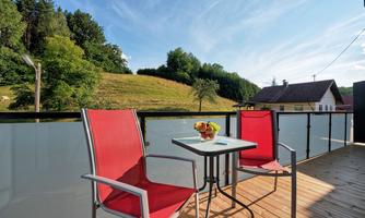 A cozy balcony with two red chairs and a small table. In the background, green hills and trees are visible.