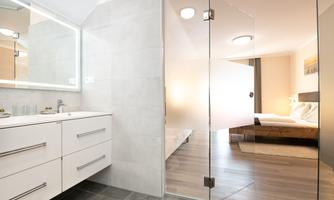 A modern bathroom with a glass shower and a white sink. In the background, a bright bedroom can be seen.