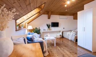 A cozy living room in the attic with wooden ceilings. It includes a sofa, a bed, and bright furniture.