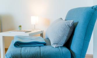 A cozy blue armchair with a cushion and a blanket. In the background, there is a small table with a lamp.