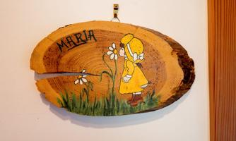 A wooden sign with the inscription "MARIA" and a childish drawing of a girl in a yellow dress, looking at flowers. The background features natural wood grain.