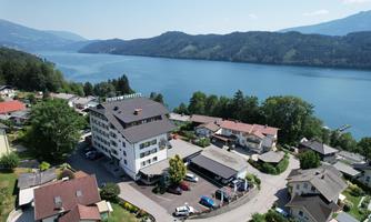 A tranquil lakeside landscape with a modern hotel in the center. Surrounded by green trees and picturesque houses.