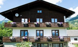 A historic house with wooden cladding and flower boxes on the balconies. The surroundings are green and mountainous.