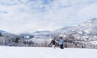A winter view of a lake surrounded by snow-covered mountains. Two people are walking hand in hand across the glittering snow surface.