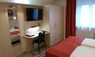 A modern hotel room with a double bed and a desk. The room has a television and large windows with red curtains.