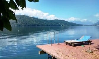 A quiet lake with a wooden pier and a lounge chair. In the background, gentle hills and a clear blue sky can be seen.