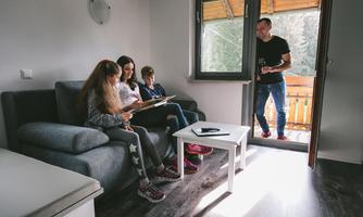 A family is sitting in the living room and looking at a tablet. A man is standing by the door and looking outside.