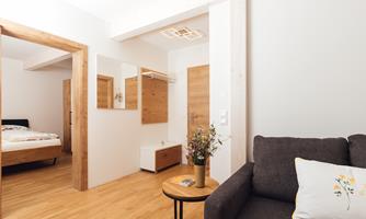 A modern living room with a cozy gray sofa and a small table. In the background, there is a sleeping area with a bed and wooden furniture.