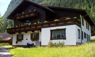An old, traditional house with a beautiful balcony, surrounded by green areas. Mountains can be seen in the background.