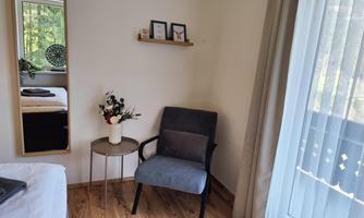 A cozy room with a chair and a small table. Pictures hang on the wall, and a large mirror reflects the light from the window side.