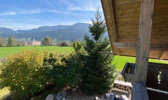 A view from a balcony onto a green meadow with mountains in the background. The landscape is sunny and clear with a blue sky.