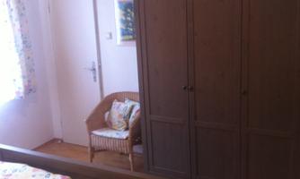 A simple bedroom with a bed and a wooden wardrobe. A chair is by the wall next to a window with a curtain.