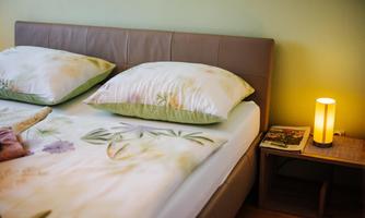 A cozy bedroom with a double bed and colorful cushions. Next to the bed is a nightstand with a table lamp and some magazines.