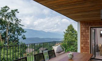 A modern balcony with a large wooden table and several chairs. In the background, a beautiful mountain landscape can be seen under a cloudy sky.