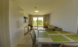 A modern living room with a dining area and a sofa. Large windows offer a view of the greenery.