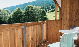 A wooden balcony with a table and chairs. In the background, green hills and forests can be seen.