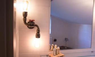A stylish bathroom lamp made of piping material emits warm light. In the background, there is a mirror and a small shelf with various bottles visible.