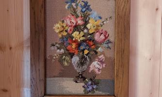 An embroidered picture with a colorful flower arrangement in a vase. The frame is made of wood and the picture hangs on a wooden wall.