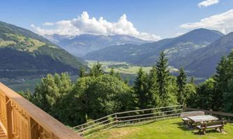 A picturesque landscape with high mountains and green forests. In the foreground, there is a wooden terrace with a table and benches.