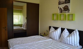 A bright bedroom with a double bed and striped bedcovers. In the background, there is a wardrobe with a mirror and a green curtain.