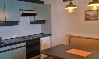 A modern kitchen with blue cabinets and a table. There is a comfortable bench at the table and there is a warm light source.