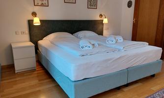 A cozy bedroom with a large double bed and fresh towels. The walls are decorated, and there is a bedside table as well as a wooden wardrobe door.