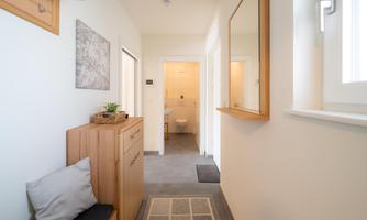 A modern hallway with light walls and a wooden cabinet. At the end of the hallway, a bathroom is visible.