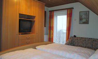 A cozy bedroom with two beds, a television, and a window with curtains. The room has a warm wooden ceiling and is simply furnished.