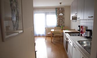A bright, modern kitchen with a dining table and chairs. Large windows provide natural light.