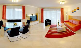 A modern living room with a red-orange sofa, a glass table, and elegant black chairs. Large windows provide plenty of daylight and a view of a colorful carpet.
