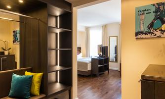 A modern hotel room area with a simple shelf, a seating area, and a television. Bright walls and large windows are visible, creating a friendly impression.