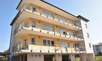 A modern residential building with several floors and balconies. The facade is yellow and white, surrounded by a clear sky.