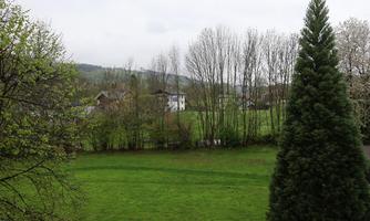 A green meadow surrounded by trees and a small house in the background. The sky is cloudy and the landscape appears calm and close to nature.