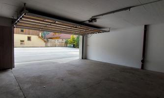 An empty garage space with an open garage door. In the background, some buildings and trees are visible.