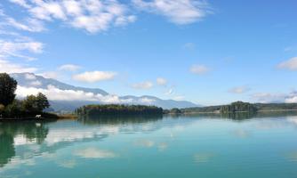 A calm lake with clear turquoise water and gentle hills in the background. The sky is blue with a few white clouds.