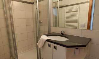 A modern bathroom with a shower and a sink cabinet. The walls are light tiled and there is a large mirror above the sink.