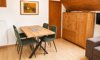 A cozy dining area with a wooden table and four chairs in soft green tones. In the background, there is a wooden piece of furniture and a painting on the wall.