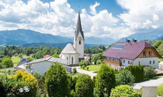 A picturesque landscape with a church tower and several houses. In the background, mountains and a blue sky with clouds can be seen.