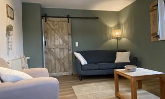 A cozy living room with a gray couch and a wooden table. On the wall is a rustic wooden door, and the lighting creates a warm atmosphere.