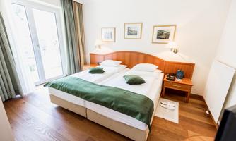 A modern hotel room with two single beds and a green bedspread. Large windows provide plenty of daylight, and there are simple furnishings for decoration.