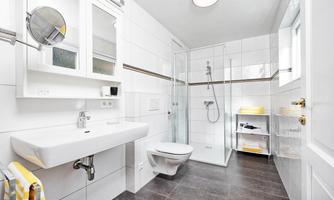A modern bathroom with white tiles and a transparent shower. There is a sink, a toilet, and an open shelf for towels.