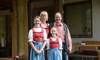 A family in traditional clothing stands in front of a wooden house. They smile warmly and pose for the photo.