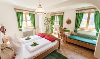 A bright bedroom with two beds, red bedding, and an additional sleeping space. Large windows with green curtains create an inviting atmosphere.